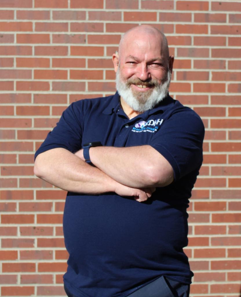 Headshot of NCDHH Outreach Liaison, Michael Brummer. Michael is a bald white man wearing a navy blue NCDHH polo shirt. Michael is standing with his arms crossed, smiling in front of a brick wall.